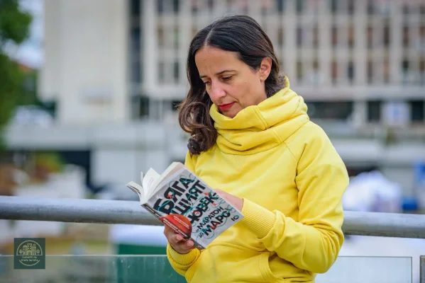 Райна Димитрова с жълт суитшърт чете отворена книга край парапет на открито.