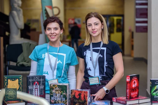 Две усмихнати жени с фестивални тениски позират зад щанд с книги.