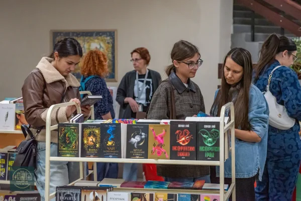 Посетителки разглеждат книги на двуетажна стойка сред книжните щандове.