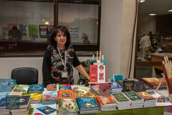 Жена зад щанд с детски книги, подредени на купчини и стойки.